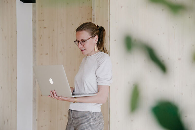 Communicatiemedewerker en copywriter Sveva leunt met laptop in de handen tegen een houten wand.
